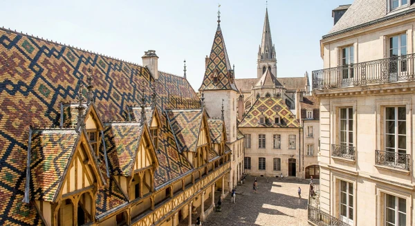 Photographie professionnelle d'une vue caractéristique de Beaune : la basilique Notre-Dame ou les toits distinctifs avec leurs tuiles vernissées colorées en motif de Bourgogne, prise depuis une perspective urbaine claire permettant d'identifier visuellement la ville. L'image doit être une photographie réaliste de qualité professionnelle, claire et accueillante, montrant l'architecture et le contexte urbain emblématique de Beaune.